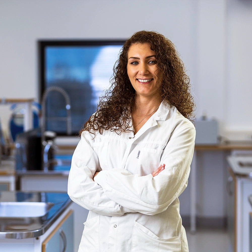 Confident female scientist in a white lab coat smiling with arms crossed in a modern laboratory setting.