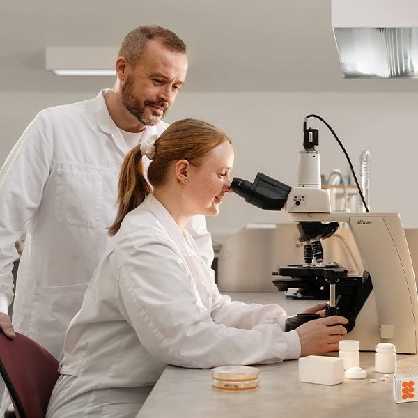 Female scientist looking into a microscope while a male colleague stands beside her in a laboratory, with lab equipment on the table
