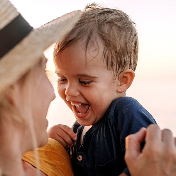 Joyful toddler laughing while being held by a woman wearing a straw hat at the beach during sunset.