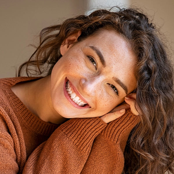 Smiling woman with curly hair and freckles wearing a warm brown sweater, radiating happiness and health.