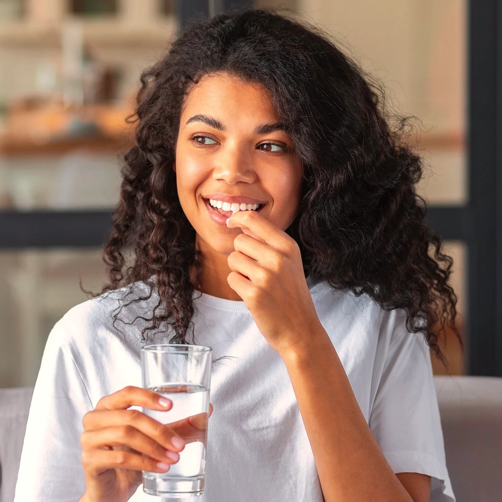 Smiling woman holding a probiotic tablet and a glass of water, about to take the supplement in a bright indoor setting.