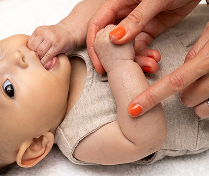 Close-up of a baby's arm held by an mother's hand with orange nail polish on a white background applying BioGaia Skincare Aldermis Probiotic Ointment.