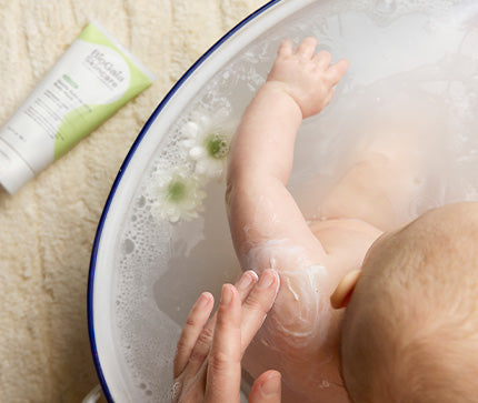 Baby taking a bath with milky water from the BioGaia Skincare Balm-to-Milk Body Wash and flowers in the water.
