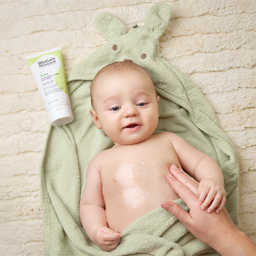 Baby wrapped in a green towel with a bottle of the BioGaia Skincare Balm-to-Milk Body Wash next to him on a textured surface with the mom's hand rubbing the balm on his body.