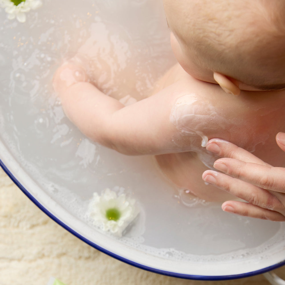 Baby taking a bath with milky water from the BioGaia Skincare Balm-to-Milk Body Wash and flowers in the water.