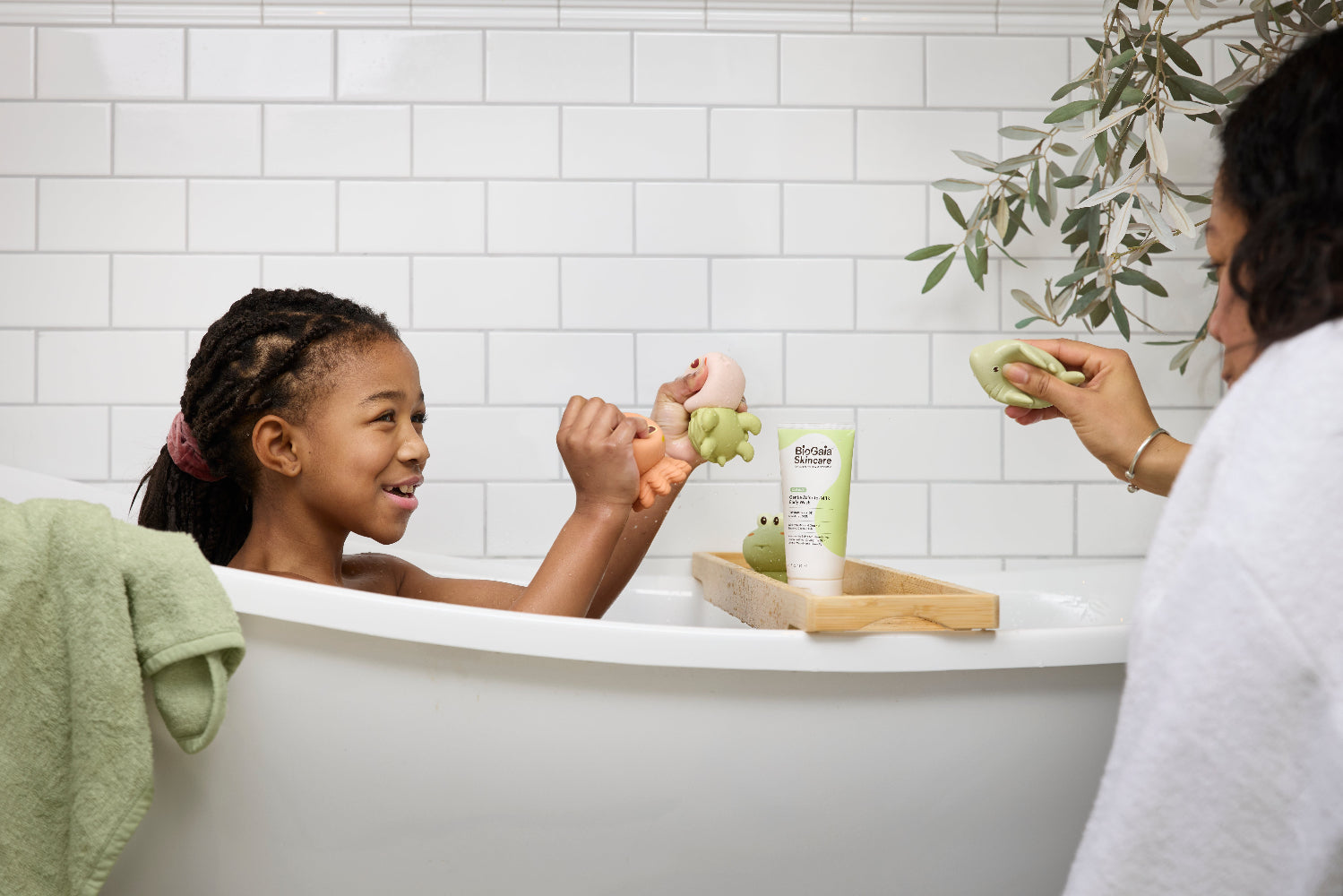 A mom and a young child enjoy a bath together in a white bathtub. They are playing together with some toys and there is the BioGaia Skincare Balm-to-Milk Wash pack on a wooden tray across the bath.