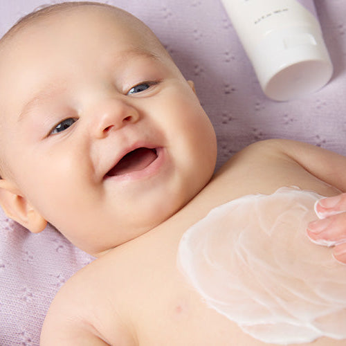 Mother's hand applying BioGaia Skincare Soothing Face & Body Lotion to her smiling baby on a lilac textured blanket background.