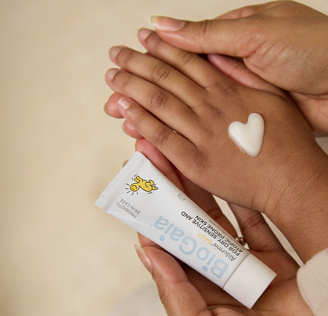 Mom and child hands holding BioGaia Aldermis Probiotic Ointment beside a heart-shaped dab of ointment on the skin. 
