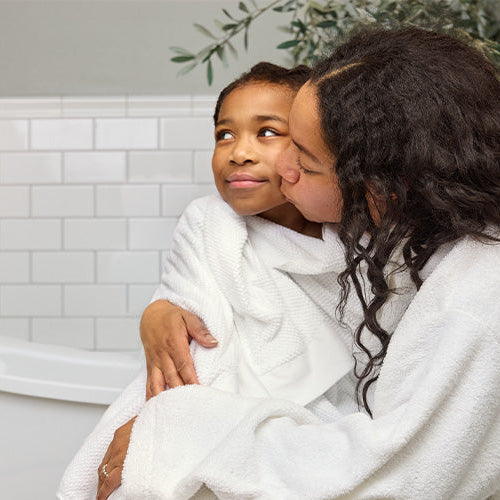 Mother and child in white robes embracing in a bathroom with mom giving the child a kiss on the cheek.