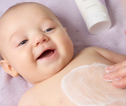 Mother's hand applying BioGaia Skincare Soothing Face & Body Lotion to her smiling baby on a lilac textured blanket background.