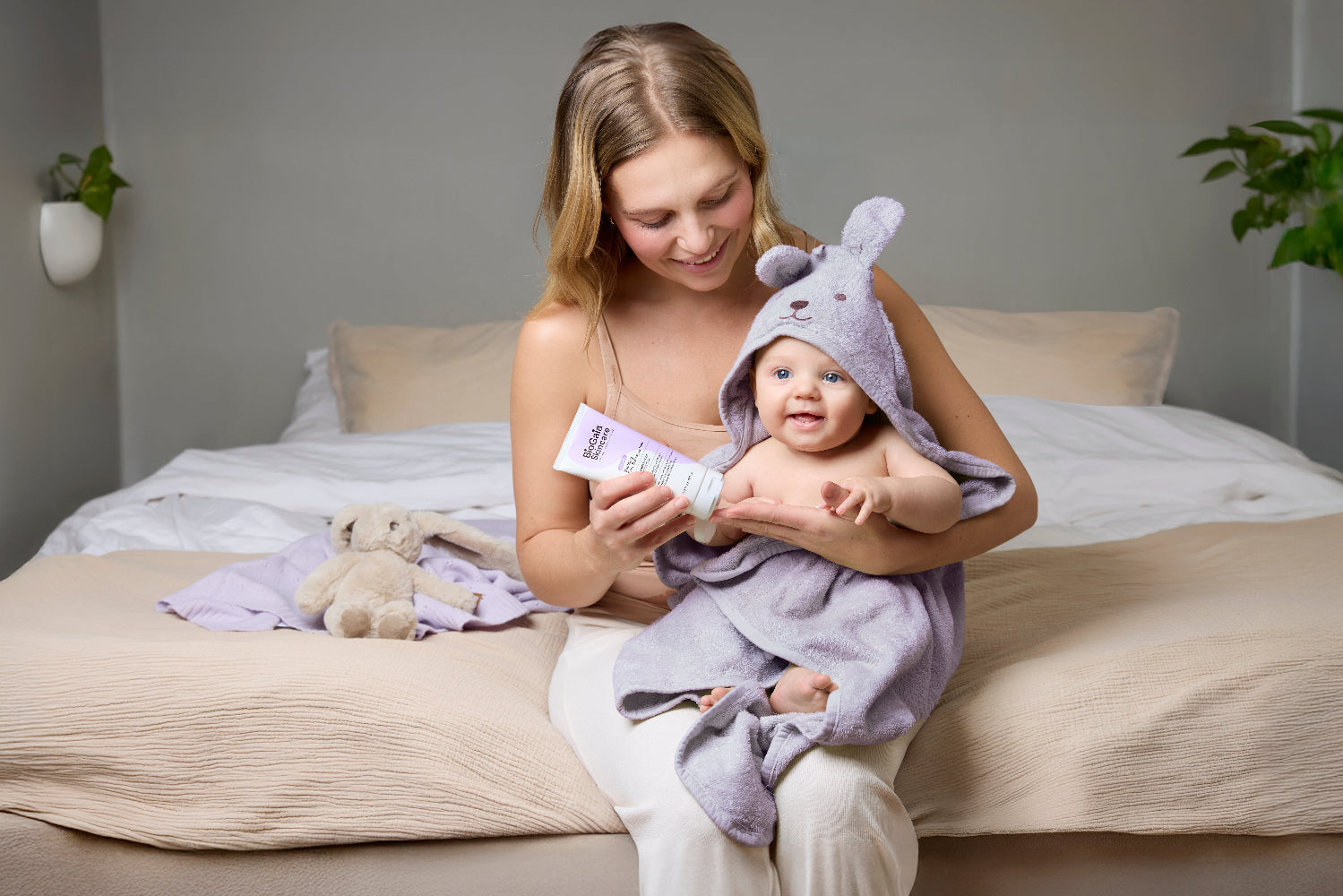 A mother sitting on a bed holds her baby wrapped in a lavender hooded towel while the woman applies BioGaia Skincare Soothing Face & Body Lotion.